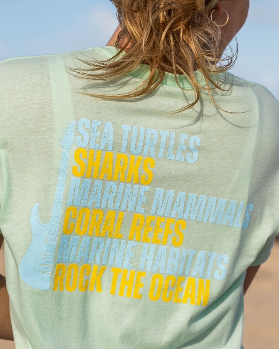 A person shown from behind wearing a light mint green graphic t-shirt. The back of the shirt features a stack of marine-themed words in light blue and yellow text ('SEA TURTLES', 'SHARKS', 'MARINE MAMMALS', 'CORAL REEF', 'MARINE HABITATS', 'ROCK THE OCEAN') next to a light blue guitar graphic.